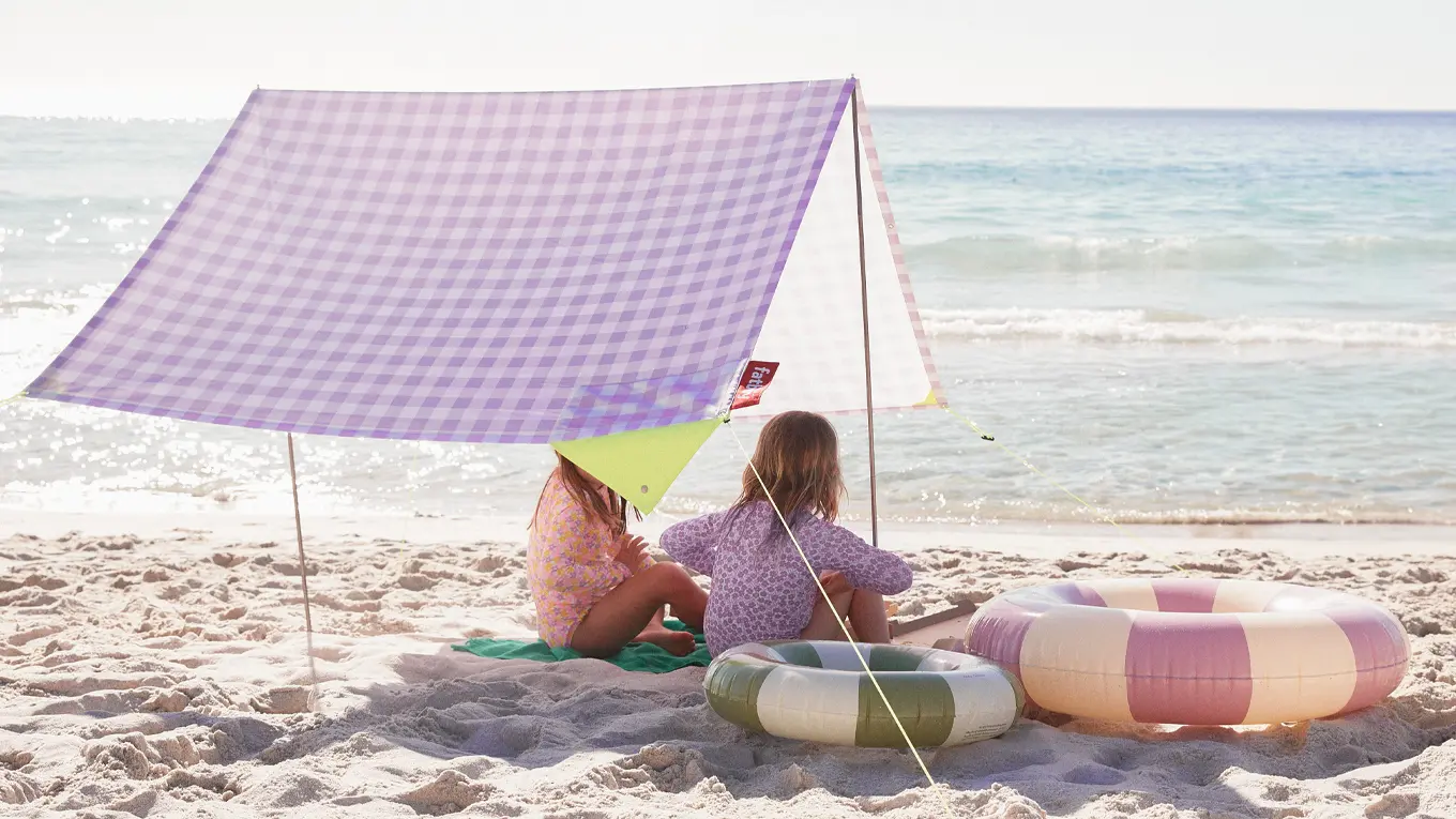 Zwei  kinder sitzen am strand unterm fatboy miasun strandzelt in lila karo